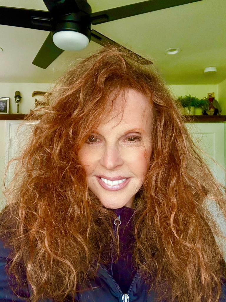 Woman with long, curly red hair smiles at the camera, wearing a dark jacket. A ceiling fan and indoor decor create a cozy atmosphere.